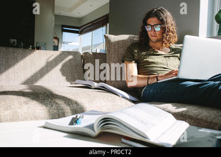 Giovane uomo studiando per gli esami seduti sul divano di casa con la luce del sole proveniente dalla finestra. Studente per esame su laptop con libri di fronte. Foto Stock