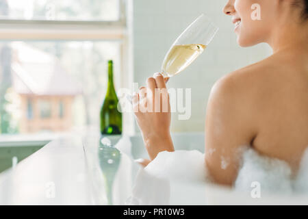 Ritagliato colpo di sorridente giovane donna bere champagne nella vasca da bagno con schiuma Foto Stock