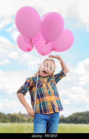 Basso angolo vista di carino bambino con palloncini rosa in campo estivo con blu cielo nuvoloso sullo sfondo Foto Stock