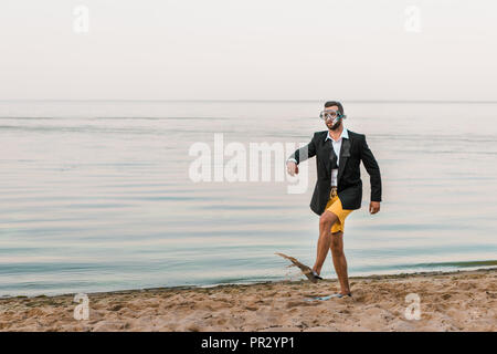 Uomo in camicia nera e shorts walking nuoto con maschera e pinne sulla riva del mare Foto Stock