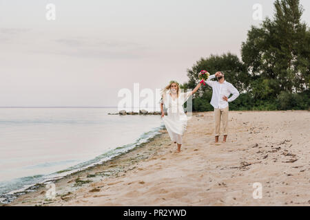 Felice sposa in esecuzione con bouquet di nozze, sposo smorfie sulla spiaggia Foto Stock