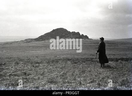 1930s, storico, una signora con un bastone in piedi di fronte Longaford Tor, un picco roccioso che si affaccia su parco nazionale di Dartmoor nel Devon, Inghilterra, Regno Unito, amn aprire selvaggio paesaggio naturale che contiene molti anicent affioramenti granitici noto come tori. Foto Stock