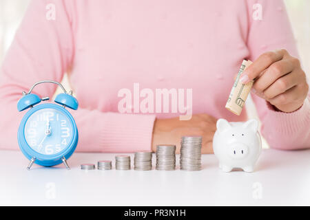 Donna mettendo mano America banconote di dollari soldi nel salvadanaio con la pila di monete e sveglia. Risparmio di denaro di ricchezza e di concetto finanziario. Foto Stock