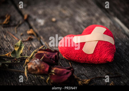 Aiuti di prima fascia imbottita e striscia di gesso Patch medico incollato sul cuore rosso con secchi rosa rossa su sfondo di legno. Cuore rotto, amore e San Valentino da Foto Stock