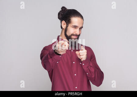 Ehi voi. Ritratto di uomo bello con scuri raccolti i capelli lunghi e la barba in maglietta rossa in piedi e puntando alla fotocamera con sorriso. piscina studio shot, Foto Stock