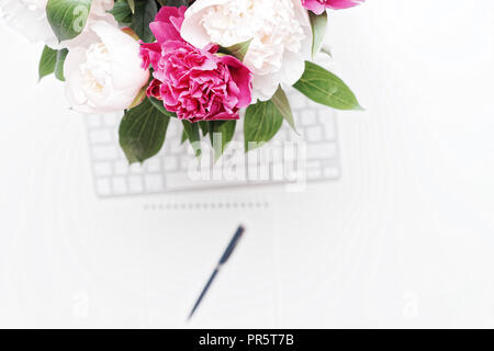 Il luogo di lavoro con tastiera, penna e bianco e rosa peonia fiori sul tavolo bianco sullo sfondo. Piatto, laici vista superiore Foto Stock