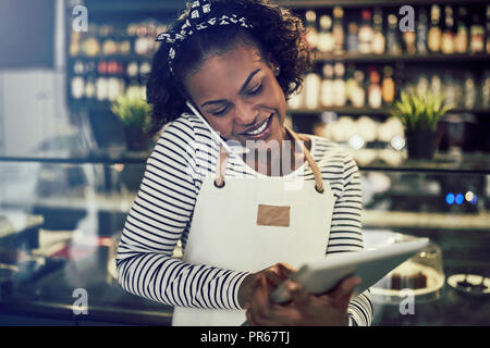 Sorridente giovane africana cafe proprietario prendendo le prenotazioni tramite il telefono e utilizzando una tavoletta digitale Foto Stock