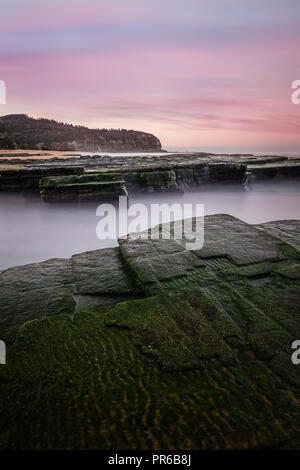 Rosa luminoso Cielo di tramonto con rocce di muschio in primo piano dalla spiaggia Turimetta. Foto Stock