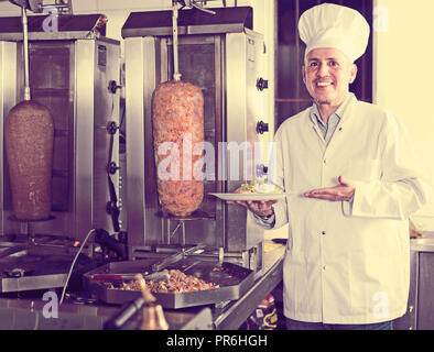 Uomo maturo cuoco indossando uniforme tenendo la piastra con carne di fresco in cafe Foto Stock