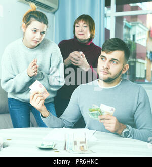 Frustrato uomo a malincuore dare contanti alla giovane moglie in piedi con la madre e mendicando più Foto Stock