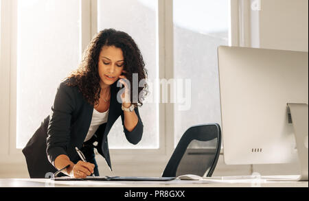 Donna imprenditore rendendo note in piedi alla sua tavola in ufficio durante la conversazione su cellulare. Imprenditrice iscritto nel diario appoggiata sulla sua tavola t Foto Stock