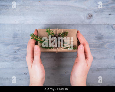 Mani femminili tenere vacanze di Natale fatti a mano presente nella carta craft con rami di abete rosso grigio su un tavolo di legno. Vista dall'alto. Sfondo di natale. Spazio di copia Foto Stock