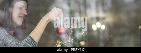 Ampia vista immagine di una giovane donna sorridente appendere rosso sfera di vacanze su un albero di Natale. Vista attraverso una finestra. Foto Stock