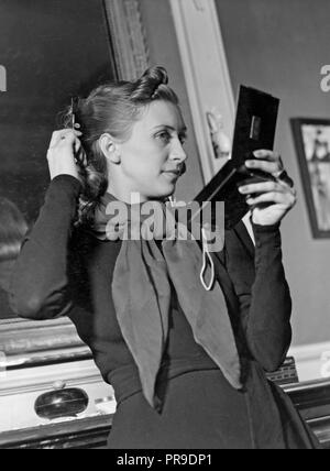 Donna pettinare i capelli. Un pelo scuro giovane donna è alla ricerca di sé in uno specchio tascabile mentre che fissa i suoi capelli. 1940s Foto Stock