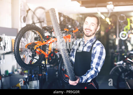 Giovane uomo in un grembiule di lavoro cambiare una ruota su una bicicletta in un negozio di biciclette Foto Stock