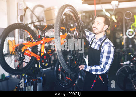 Uomo positivo nel grembiule mostra la struttura della ruota di bicicletta nel negozio di articoli sportivi Foto Stock