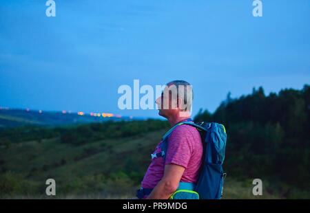 Active uomo di mezza età in piedi sulla collina con zaino, godere guardando le montagne paesaggio al crepuscolo Foto Stock
