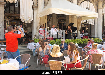 L uomo si prende la foto di orchestra che suona per i clienti di fronte iconico Florian cafe in Piazza San Marco, Venezia, Italia. Immagine mostra musicisti di suonare. Foto Stock