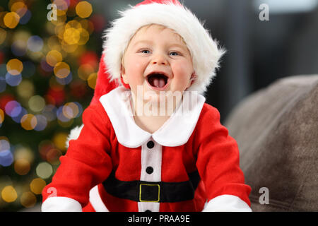Gioioso bambino indossando santa claus travestimento su un lettino a casa a Natale con un albero in background Foto Stock
