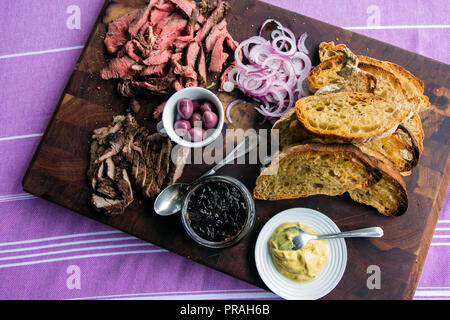 Arrosto di manzo e la bistecca sul bordo scuro con crostini di pane bianco, olive, cipolla affettata, senape e caramellato cipolla scura salsa. Foto Stock