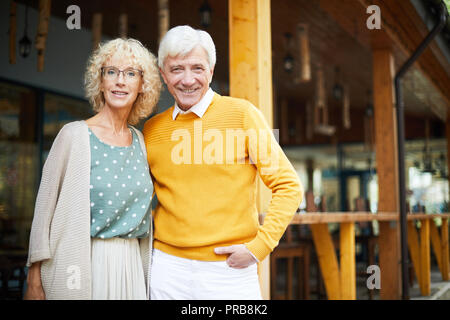 Sorridente senior alla moda giovane in piedi sulla veranda e guardando la telecamera, uomo bello che abbraccia la fidanzata in bicchieri Foto Stock