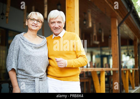 Positivo grigio bello con capelli uomo senior in maglione giallo abbracciando cara ragazza indossando abito grigio, essi in piedi sul portico e sorridente è venuto a Foto Stock