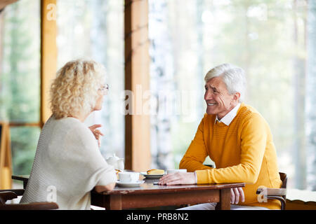 Allegro coppia senior seduta a tavola sulla veranda della loro casa di campagna e chiacchierando con piacere durante il riposo insieme nel paese Foto Stock