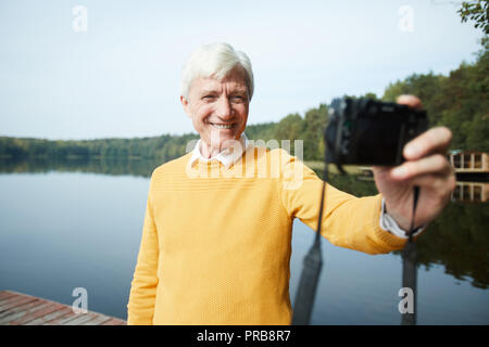 Allegro grey-haired man senior in felpa gialla essendo in vacanza in piedi sul molo e fotografare se stesso sulla fotocamera contro il lago Foto Stock