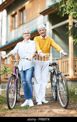 Sorridente bella coppia senior in un abbigliamento informale che conduce uno stile di vita attivo: essi azienda maniglie di bicicletta e guardando la telecamera durante il riposo di conteggio Foto Stock