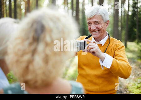 Jolly eccitato bello senior uomo in maglione giallo a fotografare gli amici sulla fotocamera e ridere mentre chiedendo loro di porre in foresta Foto Stock