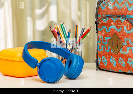 Zaino scuola con forniture scolastiche. Un supporto in metallo per matite con matite di colore giallo, casella di sandwich e cuffie su un tavolo di legno. Si torna a scuola c Foto Stock