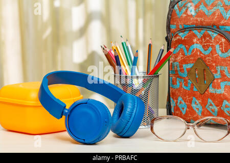 Zaino scuola con forniture scolastiche. Un supporto in metallo per matite con matite di colore giallo, casella di sandwich, cuffie e i bicchieri sul tavolo di legno. Torna alla Foto Stock