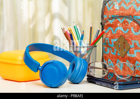 Zaino scuola con forniture scolastiche. Un supporto in metallo per matite con matite di colore giallo, casella di sandwich, cuffie, bicchieri e eBook su un tavolo di legno. Foto Stock