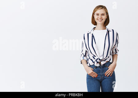 Nuovo dipendente a partire da lavoro in ufficio, sorridente durante la riunione con i colleghi di lavoro. Affascinante felice giovane donna in abiti alla moda, tenendo le mani nelle tasche e grinning dalla gioia di fotocamera, avente grande giorno Foto Stock