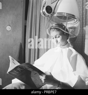 Asciugacapelli negli anni cinquanta. Una donna è comodamente seduto e la lettura rivista Vogue, mentre asciugando i capelli sotto una cappa di essiccazione. La Svezia Marzo 1954 Foto Stock