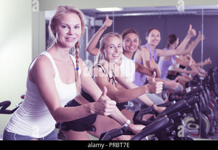 Sorridenti ragazze atletica dando pollice in alto durante l'allenamento sulla bicicletta stazionaria in palestra Foto Stock