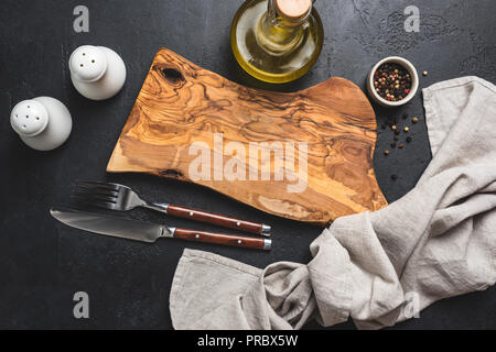 Impostazione tabella nero su sfondo di calcestruzzo. In legno di olivo tagliere, posate, sale e pepe e olio di oliva e biancheria da cucina prodotti tessili Foto Stock