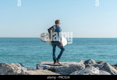 Uomo in piedi sulle rocce tenere una tavola da surf controllo del mare prima di surf. Foto Stock