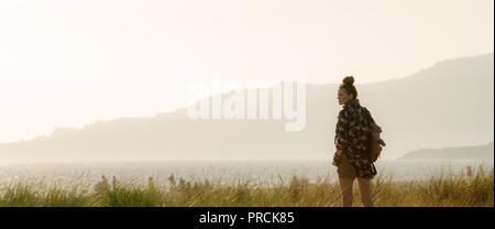 Visto da dietro il viaggiatore sano donna in abiti escursioni alla ricerca da parte contro la montagna e il mare il paesaggio al tramonto Foto Stock