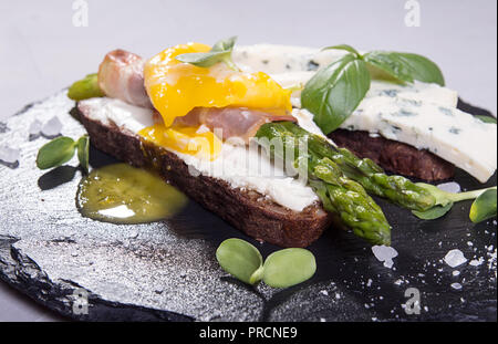 Toast con bastoni di asparagi verdi grigliati avvolto con pancetta, Uovo affogato , germogli e formaggio blu su nero ardesia pietra dello sfondo. Gray conc Foto Stock