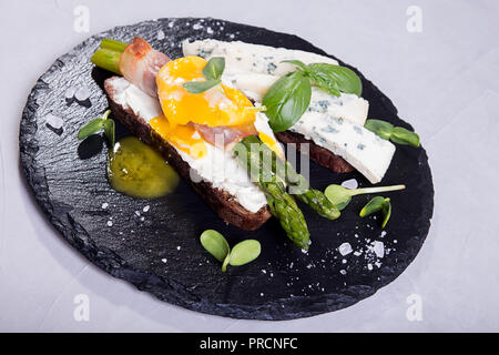 Toast con bastoni di asparagi verdi grigliati avvolto con pancetta, Uovo affogato , germogli e formaggio blu su nero ardesia pietra dello sfondo. Gray conc Foto Stock