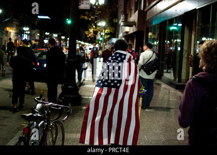Un manifestante avvolto in una bandiera americana passeggiate su Broadway in downtown Oakland durante le proteste contro la elezione di Donald Trump su nov. 9, 2016. Foto Stock