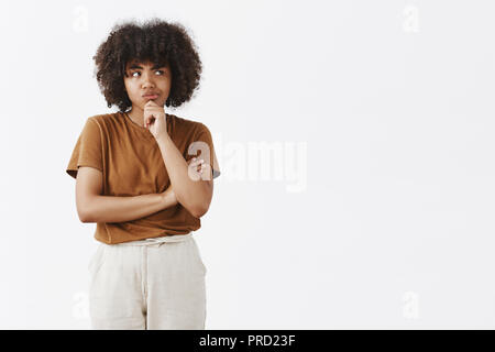 Carino di carnagione scura giovane donna intensly pensando a come risolvere la situazione. Focalizzato pensosa ragazza carina con acconciatura afro in marrone maglietta e pantaloni smirking cercando di destra mentre il pensiero di dubbi Foto Stock