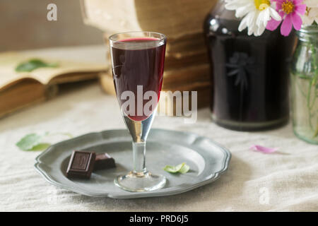 La crema de Cassis casalingo liquore di ribes nero in piccoli bicchieri, libri e fiori. Stile rustico. Foto Stock