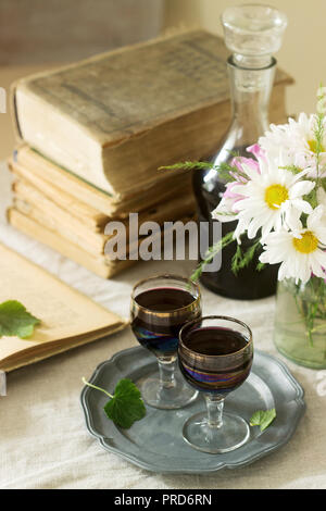 La crema de Cassis casalingo liquore di ribes nero in piccoli bicchieri, libri e fiori. Stile rustico. Foto Stock