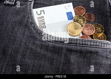 Una banconota di cinque euro e un sacco di monete si trovano nella parte anteriore della tasca dei jeans neri Foto Stock