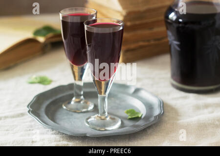 La crema de Cassis casalingo liquore di ribes nero in piccoli bicchieri, libri e fiori. Stile rustico. Foto Stock