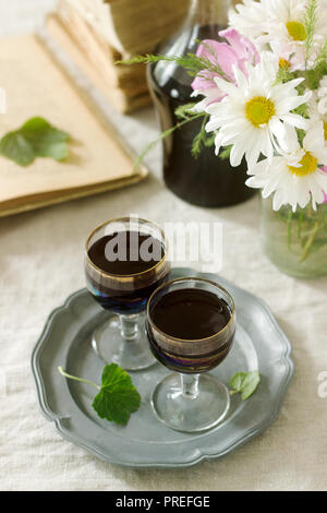 La crema de Cassis casalingo liquore di ribes nero in piccoli bicchieri, libri e fiori. Stile rustico. Foto Stock