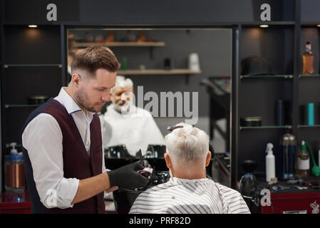 Barbiere concentrata su acconciature e rifilatura del suo cliente. La  riflessione a specchio del vecchio uomo con i capelli grigi seduto in abito  di taglio di capelli. Parrucchiere che indossa una camicia