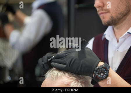 Irriconoscibile facce del barbiere e il suo cliente in barberia. Parrucchiere maschio nero che indossa i guanti mentre si lavora con i capelli. Parrucchiere il taglio di capelli grigi dell'uomo con le forbici e il pettine. Foto Stock